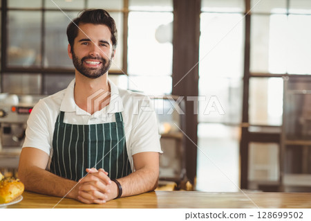Smiling male barista wearing apron behind counter in cafe with bread loaf and espresso machine 128699502