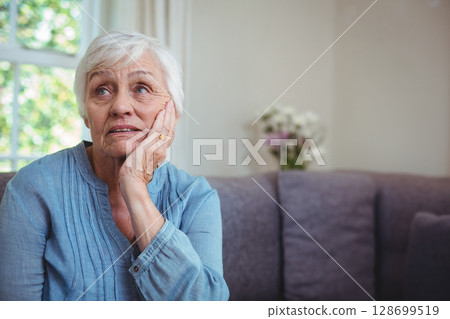 Gazing senior woman sitting on gray couch near window with white curtains, with pastel flowers 128699519