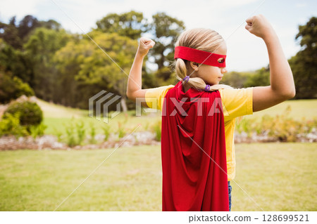 Flexing girl child posing in grassy park area near flowerbed, with red cape and mask Flexing girl child posing in grassy park area near flowerbed, with red cape and mask 128699521