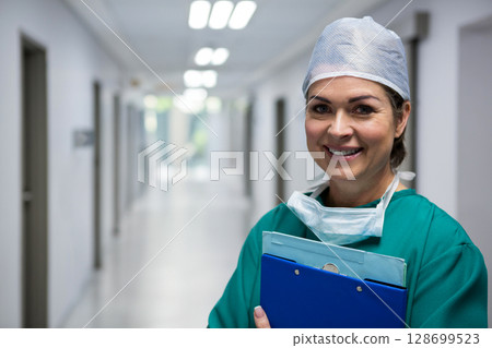 Smiling female surgeon holding blue clipboard and surgical mask in hospital hallway, copy space Smiling female surgeon holding blue clipboard and surgical mask in hospital hallway, copy space 128699523