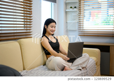 Smiling teenage girl using laptop on couch at home, enjoying online content 128699529
