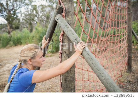 Climbing woman gripping wooden poles on forest training course, with red rope climbing net 128699533