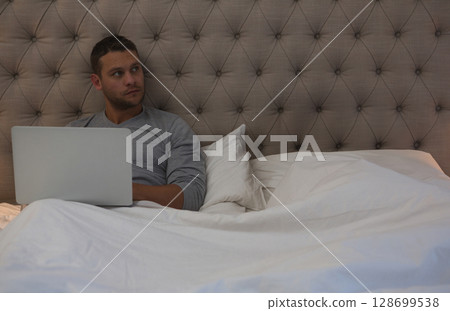 Browsing silver laptop man in his 30s sitting upright in bed in bedroom, with tufted headboard Browsing silver laptop man in his 30s sitting upright in bed in bedroom, with tufted headboard 128699538