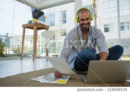 Sitting man holding printed photos on laptop in modern office, wearing neon green headphones Sitting man holding printed photos on laptop in modern office, wearing neon green headphones 128699539