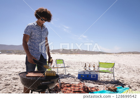 Grilling man turning corn and sausages with metal tongs on sandy beach, with cooler and chairs 128699542