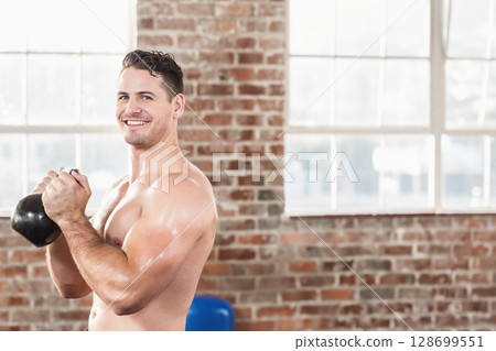 Smiling man holding kettlebell at chest height in loft gym, with blue exercise ball, copy space 128699551