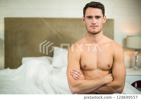 Standing man crossing arms in bedroom by upholstered headboard and crystal table lamp, copy space Standing man crossing arms in bedroom by upholstered headboard and crystal table lamp, copy space 128699557