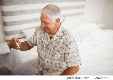 Smiling senior man holding digital thermometer while sitting in bright bedroom, copy space Smiling senior man holding digital thermometer while sitting in bright bedroom, copy space 128699559