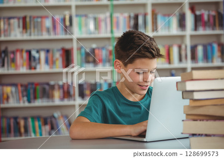 Child typing on silver laptop at library study table, with stack of hardcover books 128699573