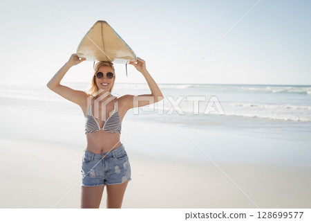 Holding surfboard overhead woman standing on sandy beach at water's edge, wearing round sunglasses Holding surfboard overhead woman standing on sandy beach at water's edge, wearing round sunglasses 128699577