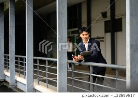 Asian businessman holding laptop leaning on railing in modern outdoor corridor 128699617