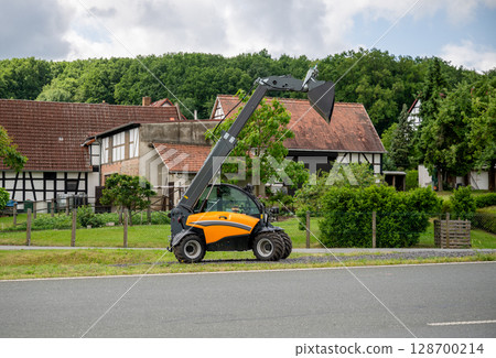 Telescopic loader with raised bucket parked near traditional countryside houses in summer Telescopic loader with raised bucket parked near traditional countryside houses in summer 128700214