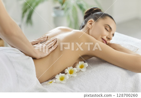 Young woman enjoying relaxing soothing back massage in spa, breathing aroma from flowers Young woman enjoying relaxing soothing back massage in spa, breathing aroma from flowers 128700260