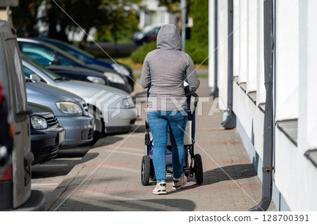 a woman with a baby carriage walks on the sidewalk along the side of the street, rear view 128700391