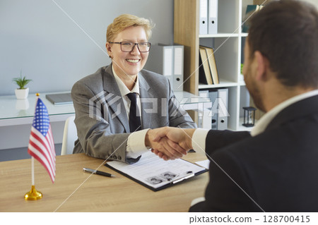 Shaking hands across desk, people sitting greeting in office near American flag, woman smiling Shaking hands across desk, people sitting greeting in office near American flag, woman smiling 128700415