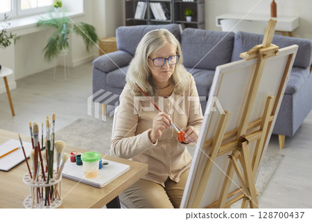 Senior artist sitting at the desk in front of easel in her workshop, making picture 128700437