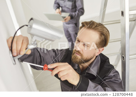 Young technician man in uniform installing surveillance CCTV camera on the wall indoors Young technician man in uniform installing surveillance CCTV camera on the wall indoors 128700448