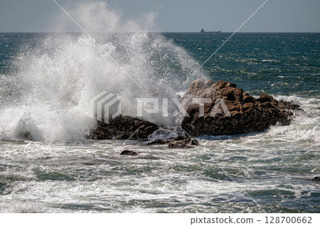 Powerful ocean waves collide with large rocks along the coast under a clear sky. Powerful ocean waves collide with large rocks along the coast under a clear sky. 128700662