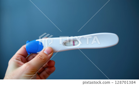 Hand holding blue and white pregnancy test with two lines indicating result, set against blue background 128701384