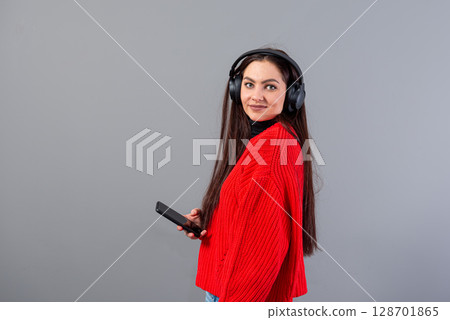 positive girl brunette in red sweater listens to music on smartphone in headphones, isolated on gray positive girl brunette in red sweater listens to music on smartphone in headphones, isolated on gray 128701865