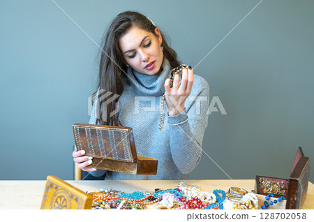 young brunette sitting at the table with lots of jewelry, choosing and trying different jewelry 128702058