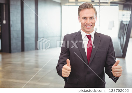 Smiling middle-aged man standing in modern office lobby, giving thumbs up with red tie, copy space 128702093