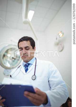 Reviewing blue clipboard male doctor wearing stethoscope in exam room, with surgical lamp and clock Reviewing blue clipboard male doctor wearing stethoscope in exam room, with surgical lamp and clock 128702099