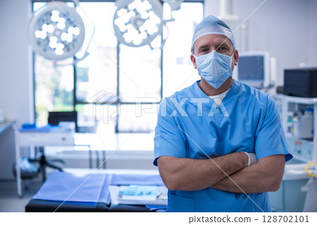 Standing male surgeon crossing arms in OR with surgical lights and blue drape, copy space Standing male surgeon crossing arms in OR with surgical lights and blue drape, copy space 128702101