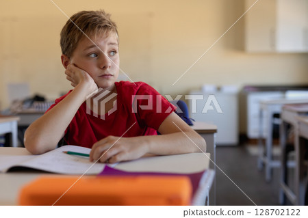 Gazing child resting cheek on hand at classroom desk, with notebook and pencil, copy space 128702122