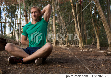 Man performing seated shoulder stretch on forest trail, with wooden rail and soft sunlight Man performing seated shoulder stretch on forest trail, with wooden rail and soft sunlight 128702123
