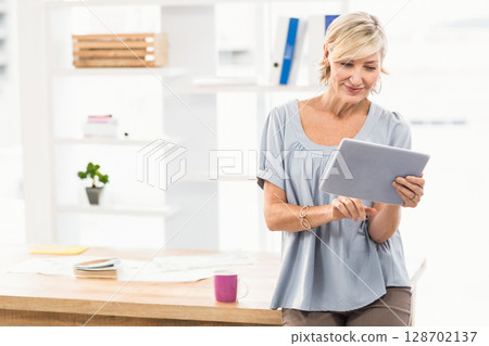 Woman leaning against desk tapping tablet in office, with papers, coffee mug, copy space 128702137