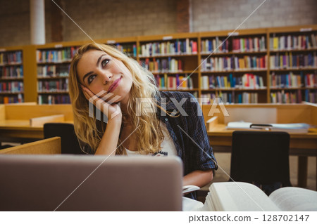 Gazing woman sitting at modern library table, with open laptop and thick textbook 128702147