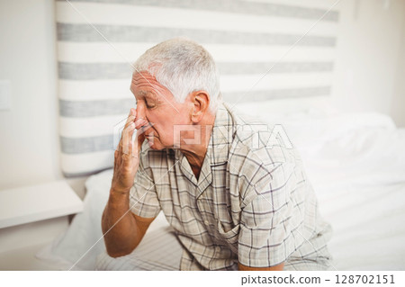 Rubbing face Senior man sitting on bed edge in bedroom, with striped headboard, bedside table Rubbing face Senior man sitting on bed edge in bedroom, with striped headboard, bedside table 128702151