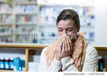 Sneezing woman holding tissue at wooden pharmacy counter, with blurred medicine shelves, copy space 128702160