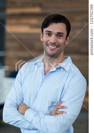 Man standing with crossed arms and smiling in modern office interior, with wooden accent wall 128702166