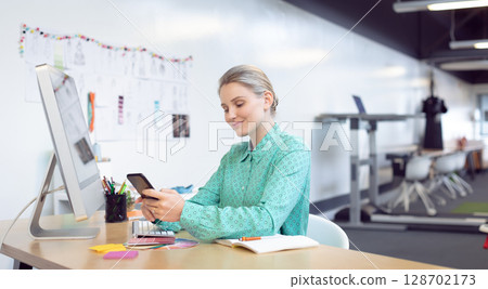 woman scrolling smartphone at desk in open office, with computer monitor and swatches woman scrolling smartphone at desk in open office, with computer monitor and swatches 128702173