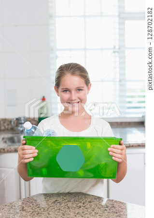 Holding green recycling bin, female child standing behind kitchen counter showing plastic bottles 128702182