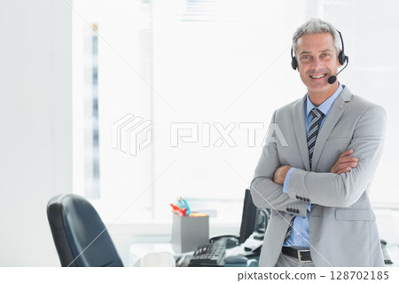 Smiling man standing in corporate office wearing headset with monitor and keyboard, copy space 128702185
