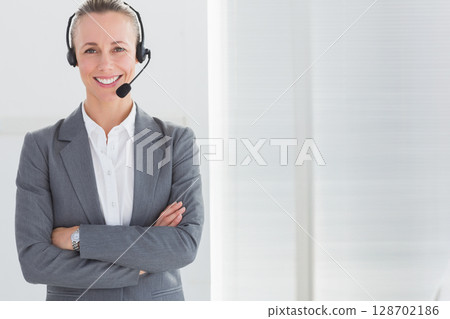Smiling woman wearing headset and watch crossing arms standing in office near blinds, copy space 128702186