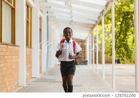 Walking African American school boy carrying red backpack along covered outdoor corridor, sunny day 128702200