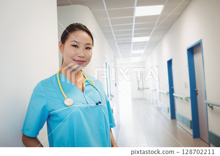Leaning Asian woman wearing blue scrubs in hospital corridor, with yellow stethoscope, copy space 128702211