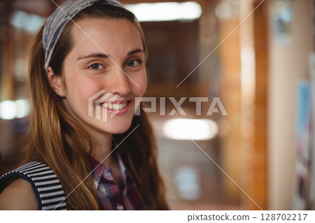 Smiling teenage girl standing in school corridor, holding striped backpack and patterned headband 128702217