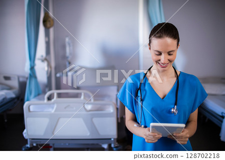 Smiling female nurse reviewing tablet with stethoscope in hospital ward, copy space 128702218