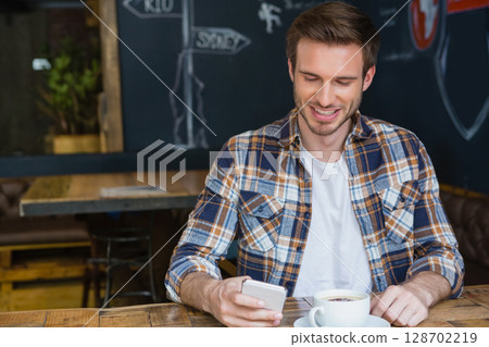 Smiling man sitting at rustic cafe table holding smartphone with coffee cup on saucer, copy space 128702219