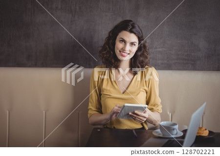 Smiling woman holding tablet while sitting at wooden cafe table with laptop, coffee and croissant 128702221