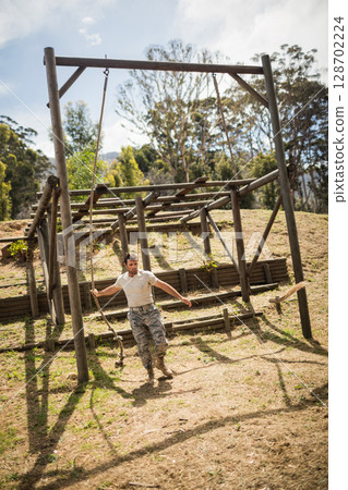 Climbing man navigating obstacle frame on wooded hillside, with hanging rope and log barriers Climbing man navigating obstacle frame on wooded hillside, with hanging rope and log barriers 128702224