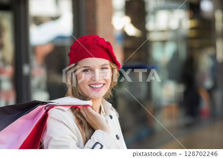 Smiling woman carrying colorful paper shopping bags in shopping district, with red knit beanie 128702246