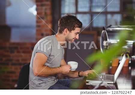Man typing on silver laptop at industrial-style coffee shop counter, holding coffee mug 128702247