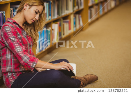 Sitting woman reading open book on lap in library aisle, leaning against wooden bookshelf Sitting woman reading open book on lap in library aisle, leaning against wooden bookshelf 128702251