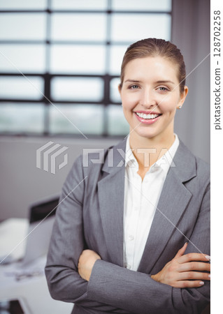 Smiling woman standing with arms crossed in office, with computer monitor and files, copy space Smiling woman standing with arms crossed in office, with computer monitor and files, copy space 128702258